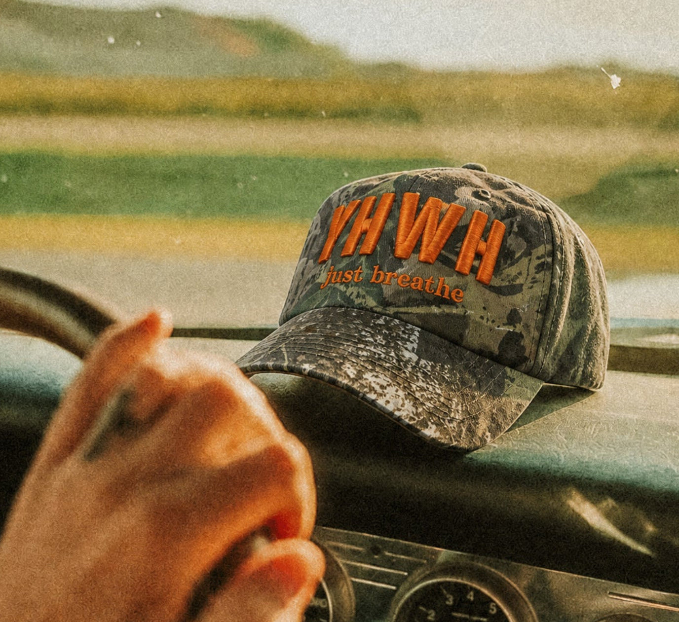 A camo baseball cap with orange embroidered text reading 'YHWH' and sits on a car dashboard while a hand rests on the steering wheel, with a blurred outdoor landscape visible through the windshield.